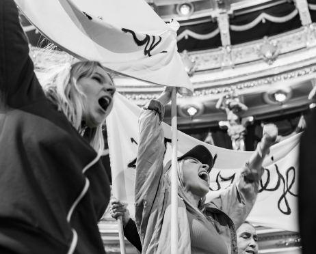 Czarno-białe zdjęcie. Na pierwszym planie Karolina Kamińska i Karolina Kazoń we wnętrzu teatru. Aktorki trzymają baner na długich drążkach, unoszą ręce i krzyczą, scena wygląda jak protest. W tle widoczne są balkony, kolumny i oświetlenie przestrzeni teatralnej.